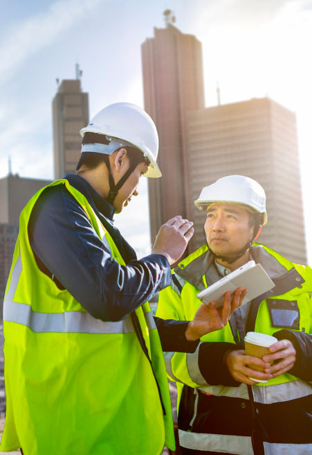Two men in hi-vis jackets have a conversation on a building site while looking at a tablet.